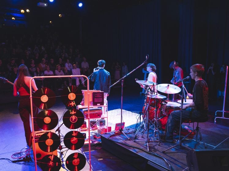 Image of A band performs on stage in front of an audience. The view is from behind the band, showing a drummer, guitarists, a rack with vinyl records, and stage equipment under blue and purple lighting.