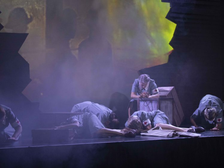 Image of Five people dressed as nurses in vintage uniforms are on stage, some kneeling and some lying on the ground, surrounded by dramatic lighting, mist, and shadowy figures in the background. The scene appears tense and emotional.