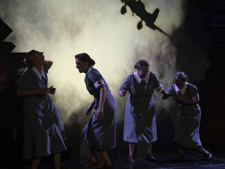 Image of Five women in Second World War–style nurse uniforms react in fear as the shadow of an aeroplane appears on a smoky, backlit stage set.