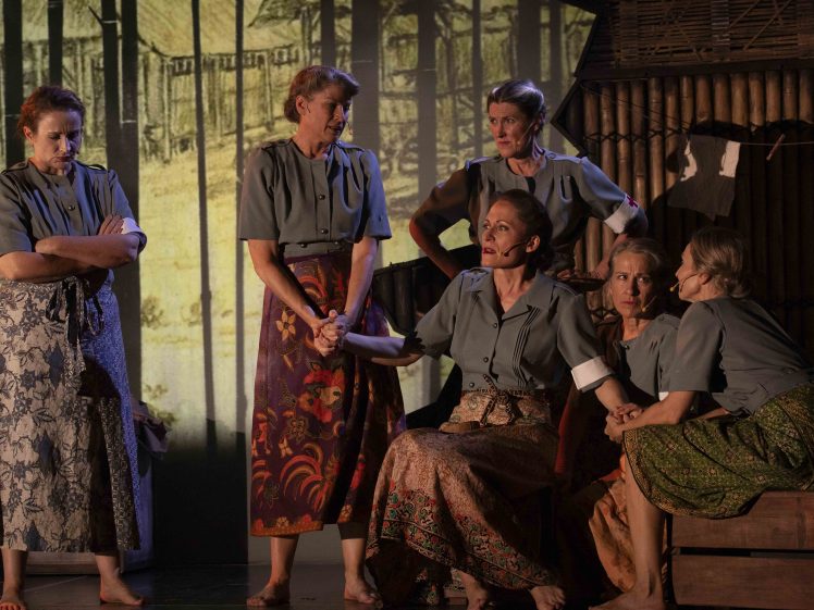 Image of Six women in grey blouses and patterned skirts are gathered on stage, some sitting and some standing, with serious expressions, in front of a backdrop showing wooden structures and trees.