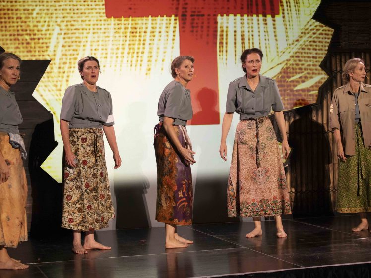 Image of Five barefoot women in grey tops and patterned skirts stand on a stage, looking concerned. A large red cross is displayed on a backdrop behind them. The setting appears to be a dramatic theatre performance.