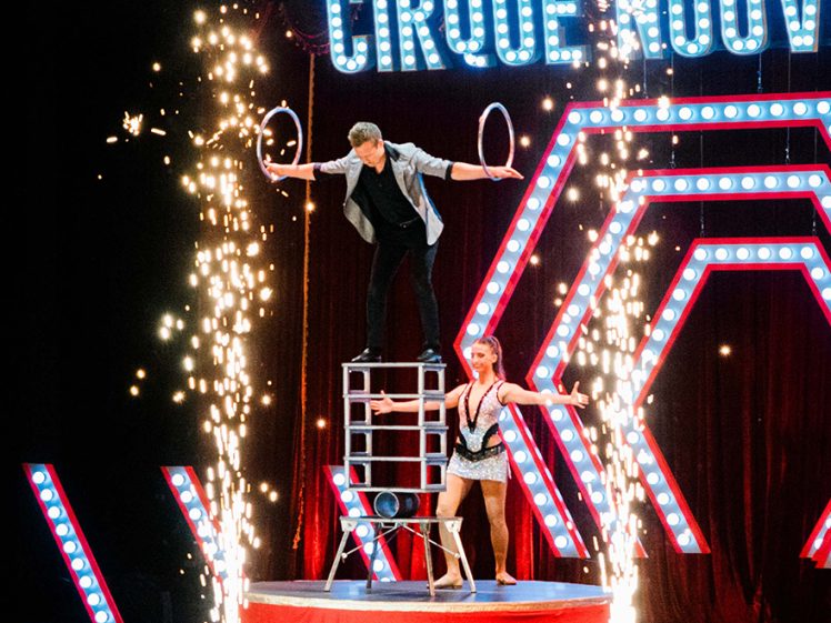 Image of Two circus performers on stage: one balancing on chairs holding hoops, the other standing on the platform below. Bright lights, sparkling pyrotechnics, and a sign reading CIRQUE NOUVEAU are in the background.