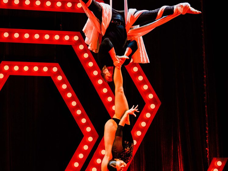 Image of Two acrobats perform an aerial routine; one hangs upside down from fabric, supporting the other by her foot. They are on a stage with bright red hexagonal lights and large illuminated letters in the background.