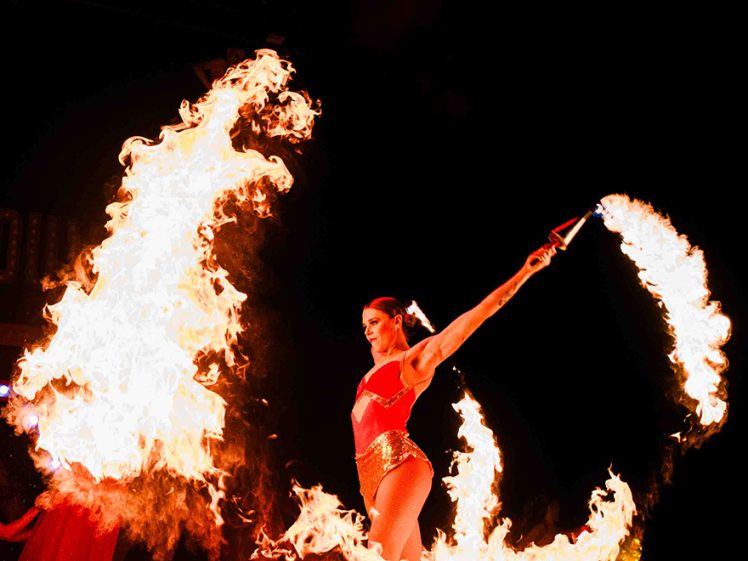 Image of A performer in a red costume spins flaming batons on a dark stage, creating large arcs of fire around her.