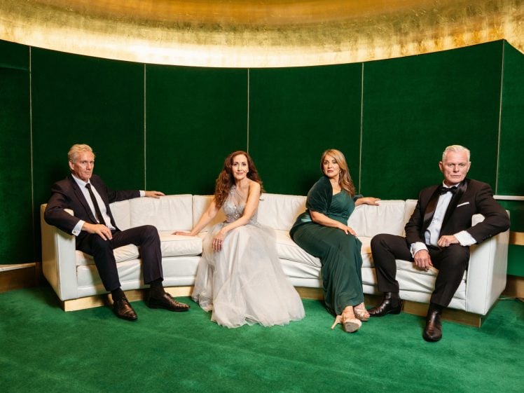 Image of Four elegantly dressed adults sit on a white sofa in a green and gold room; two men in black dinner jackets and two women in evening dresses, one silver and one green, all facing the camera.