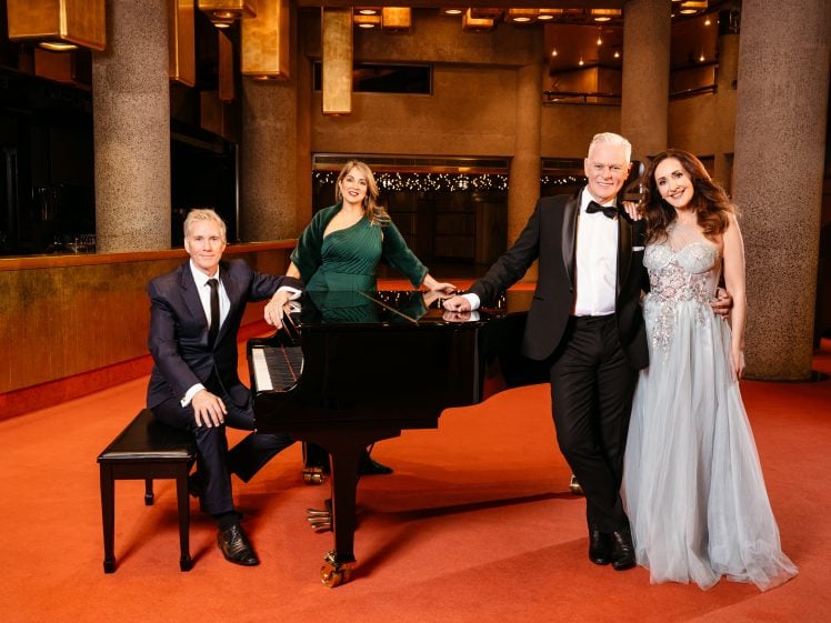 Image of Four elegantly dressed people pose around a black grand piano in a modern, warmly lit venue with orange carpet and cylindrical columns. Two women stand, one in green and one in blue, while two men in suits are at the piano.