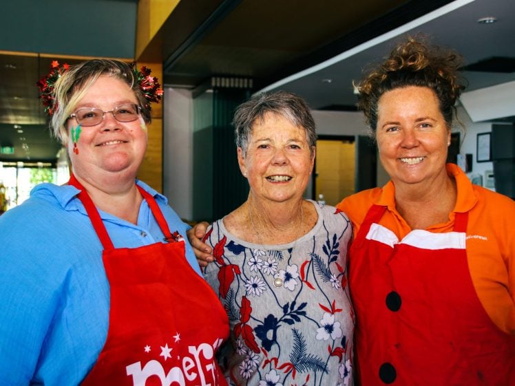 Image of Three women smiling together indoors; one wears a red apron and festive hair accessories, another wears a patterned blouse, and the third wears an orange shirt and red apron with a white collar and black buttons.