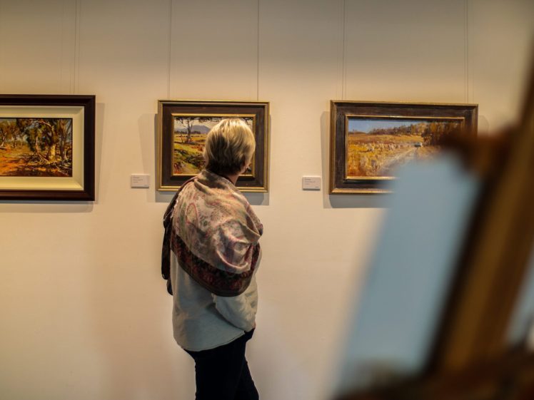 Image of A person stands in an art gallery, closely viewing framed landscape paintings on a white wall. The individual has short, fair hair and is draped in a patterned shawl. An easel is visible in the foreground.