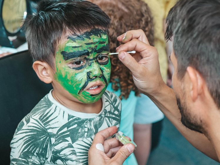 Image of A young boy with green face paint is having his face painted by an adult. He looks slightly uncomfortable and is wearing a leafy-patterned shirt. The adult is carefully detailing around his eyes.