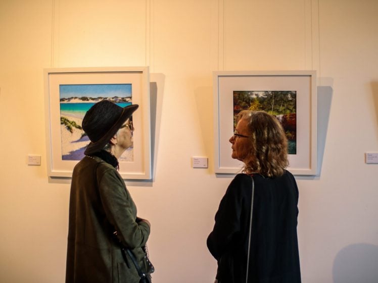 Image of Two women stand side by side in front of framed landscape paintings in an art gallery, engaged in conversation whilst viewing the artwork on a white wall.
