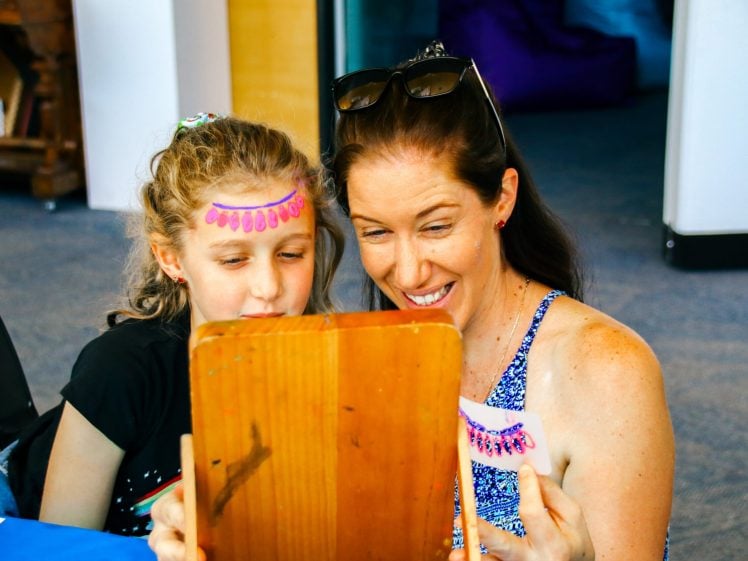 Image of A woman and a young girl smile whilst looking into a wooden mirror. The girl has a colourful decorative design on her forehead and the woman holds a matching sheet with similar designs. They appear to be enjoying a fun activity together.