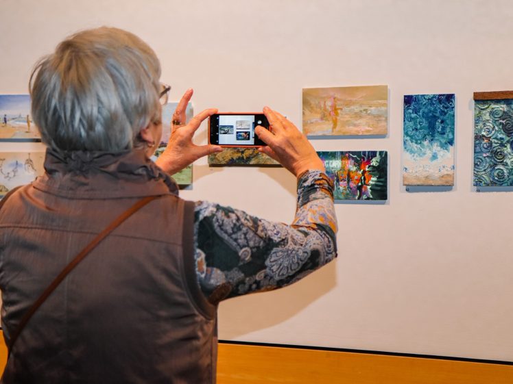 Image of An older adult with short grey hair takes a photo of small colourful paintings displayed on a gallery wall using a mobile phone.