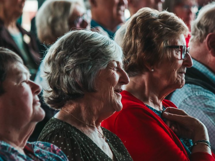 Image of A group of older adults sit closely together in an audience, smiling and engaged as they watch something off-camera. The atmosphere appears warm and lively.