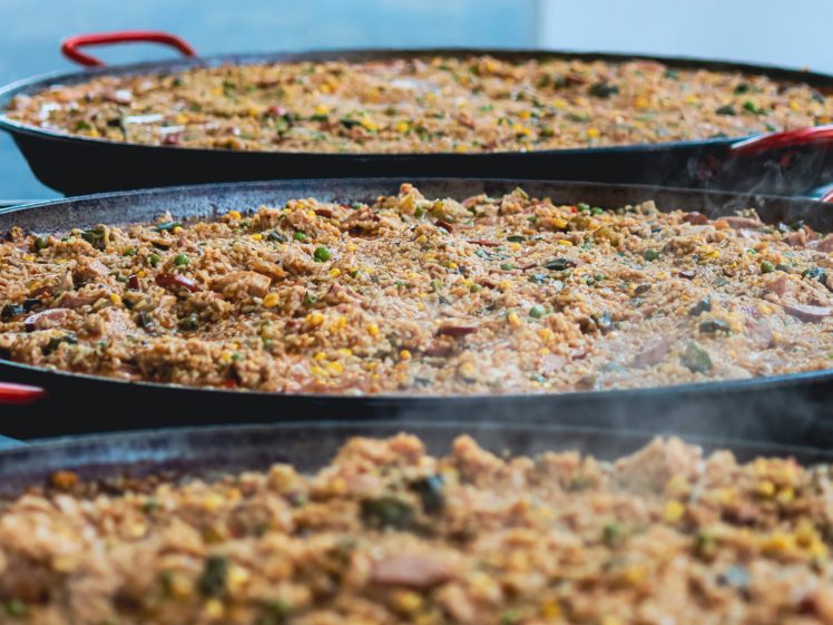 Image of Large pans filled with steaming, colourful paella packed with rice, vegetables, and various ingredients, displayed in a row with red-handled pans, ready to be served.
