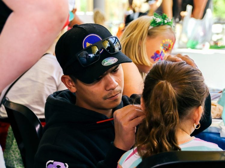 Image of A man wearing a black cap and hoodie carefully paints a design on a young girl's face at an indoor event, while other children with face paint sit nearby.