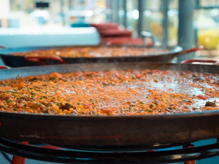 Image of Large shallow pans filled with simmering paella, featuring rice, vegetables, and mixed ingredients, sit on hobs in a bright, modern setting with glass windows in the background.