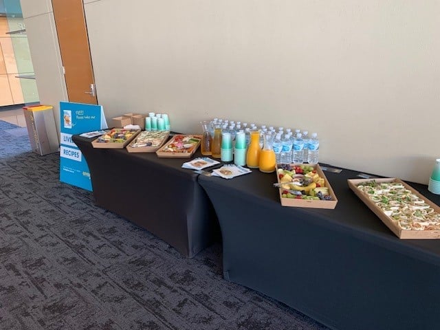 Image of A long black-covered table set with trays of assorted snacks, fruit, water bottles, juice bottles, and small turquoise cups against a beige wall at an indoor event. A blue promotional sign stands at the end of the table.