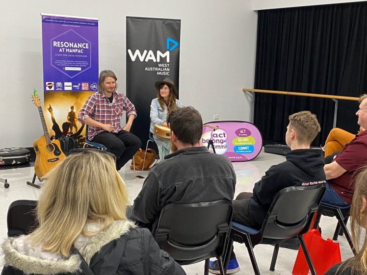 Image of A man and woman sit at the front of a small group, leading a music workshop. Guitars rest nearby, and banners for Resonance at Nannup and West Australian Music are behind them. Attendees face forwards, listening.