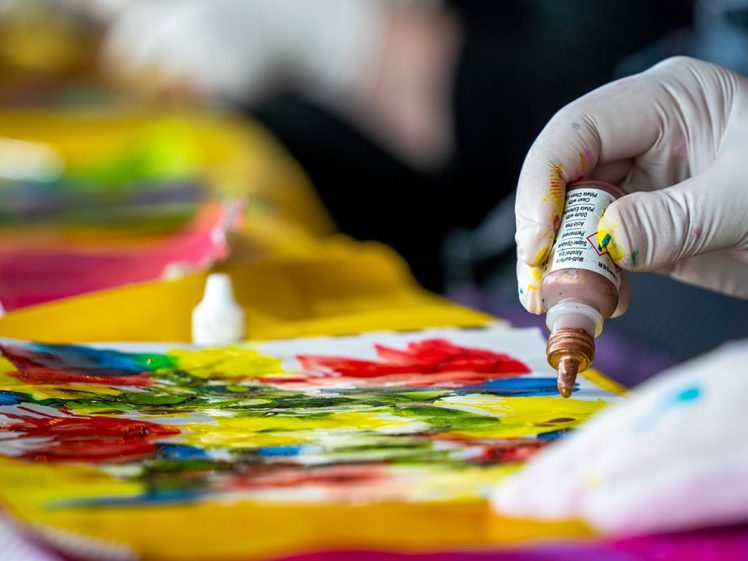 Image of A gloved hand squeezes a bottle of brown paint onto a colourful abstract artwork featuring vibrant reds, greens, and yellows on paper. Art supplies and blurred backgrounds are visible.