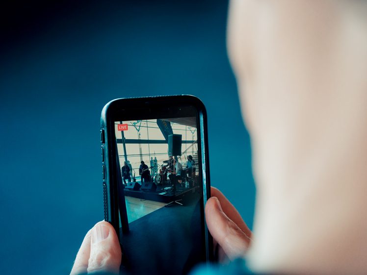 Image of A person holds a mobile phone, live-streaming a band performing on a stage with musical instruments, seen clearly on the phone screen while the foreground is out of focus.