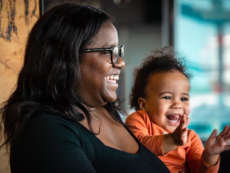 Image of A woman with glasses smiles whilst holding a happy baby in her arms. The baby, wearing an orange shirt, is laughing and clapping hands. Both appear joyful and are looking to the right.