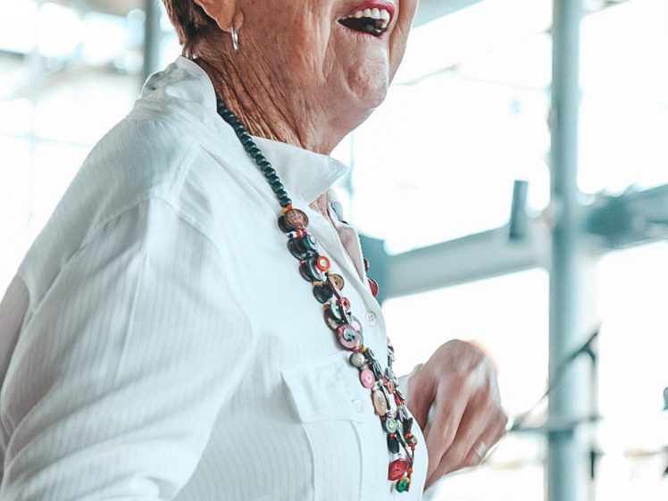 Image of An older woman with short, fair hair smiles and gestures with her hands. She wears a white blouse, a beaded necklace, and a bracelet, standing in a bright room with large windows in the background.