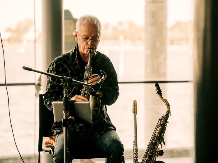 Image of An older man sits indoors playing a soprano saxophone, with a tenor saxophone resting nearby. He reads sheet music from a digital tablet, and large windows reveal a blurred outdoor view behind him.