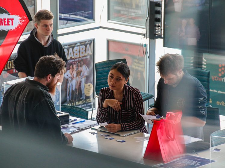 Image of Four people interact at a registration table with name badges and paperwork; a large red bag sits on the counter. Posters and windows are visible in the background.