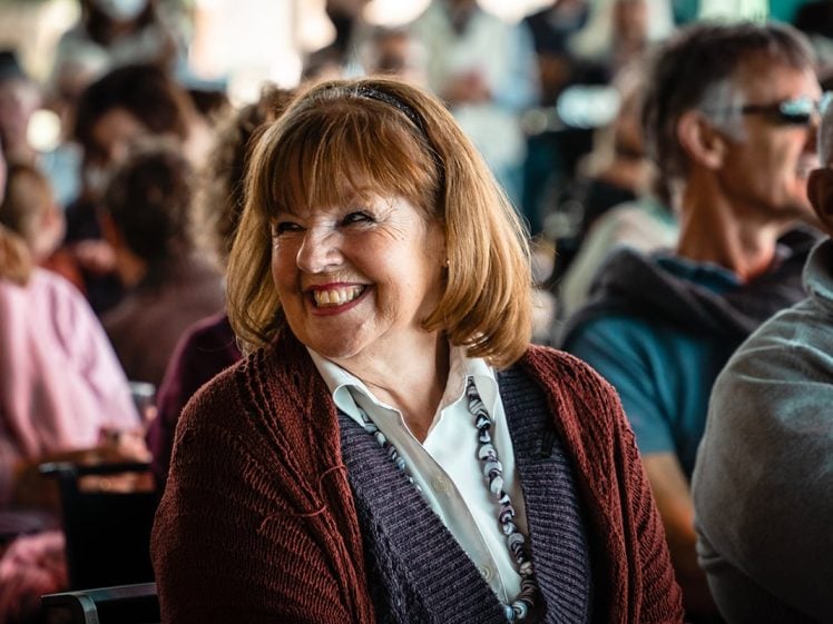 Image of A woman with red hair and a maroon jumper smiles brightly whilst sitting among a group of people at an indoor event. The background shows other attendees, some blurred and casually dressed.