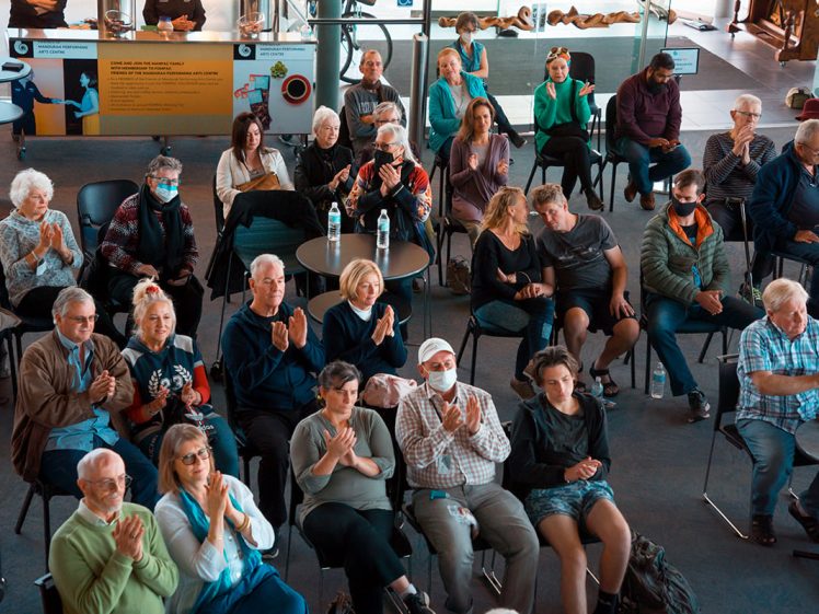 Image of A group of people sit indoors, some wearing masks, applauding as they face a presentation or performance. The audience includes men and women of various ages, seated in rows of chairs around tables.