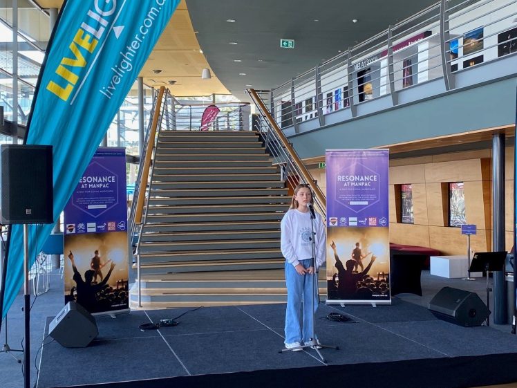 Image of A young person stands on a small stage with a microphone in front of a staircase. Banners read Livelighter and Resonance. Colourful beanbags are in the foreground and people are seated watching the performance.