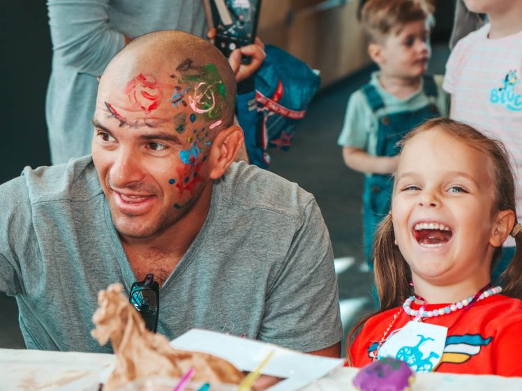 Image of A smiling man with colourful face paint sits beside a laughing young girl at a table with art supplies, surrounded by other children in a lively indoor setting.