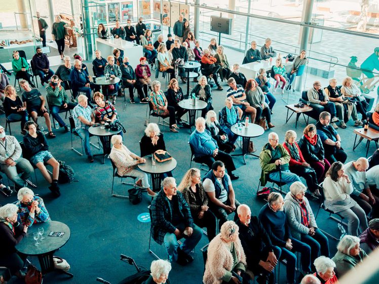 Image of A large group of people, mostly pensioners, sit at round tables in a bright, modern indoor space with glass walls, attentively facing the same direction, suggesting an event or presentation.