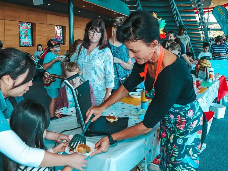 Image of A woman in a floral apron serves food to a child at a busy indoor event; people, including children, stand around decorated tables under bright lighting by a staircase.