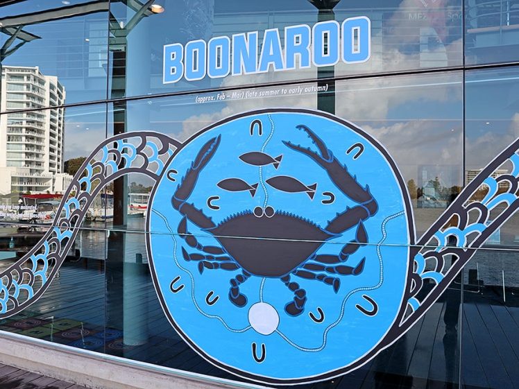 Image of Large blue mural of a crab and three fish on a glass building facade with the word BOONAROO above it; river, modern buildings, and reflections visible in the background.