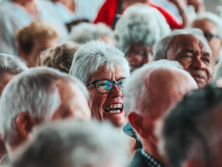 Image of A group of older adults with grey hair sit closely together in an audience. One woman in the centre, wearing glasses, is smiling and appears to be laughing. The atmosphere looks lively and joyful.