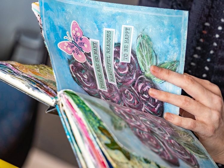 Image of A person holds an open, colourful art journal featuring painted flowers, a pink butterfly, and the text There are so many beautiful reasons to be happy.