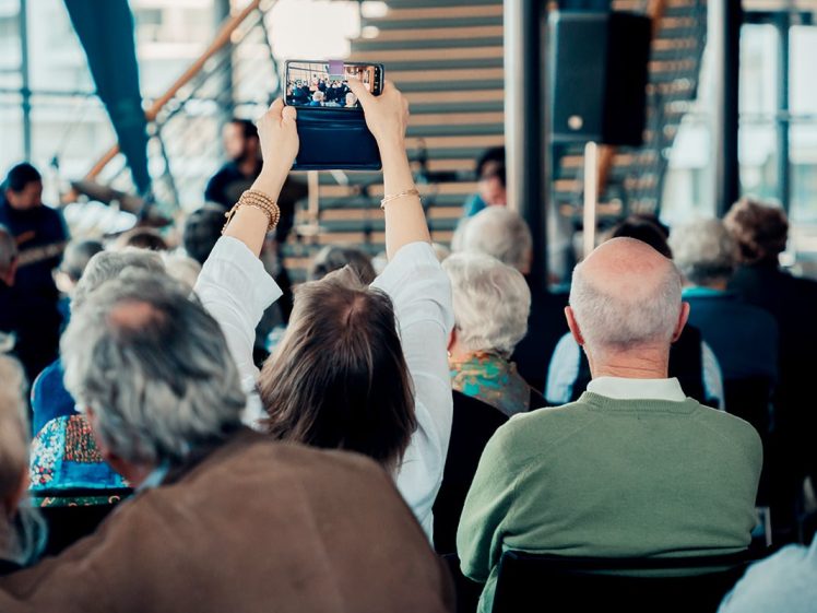 Image of An audience of mostly older adults sits facing a stage. A person in the centre raises a tablet, taking a photo or video of the event. The atmosphere suggests a public gathering or live performance.