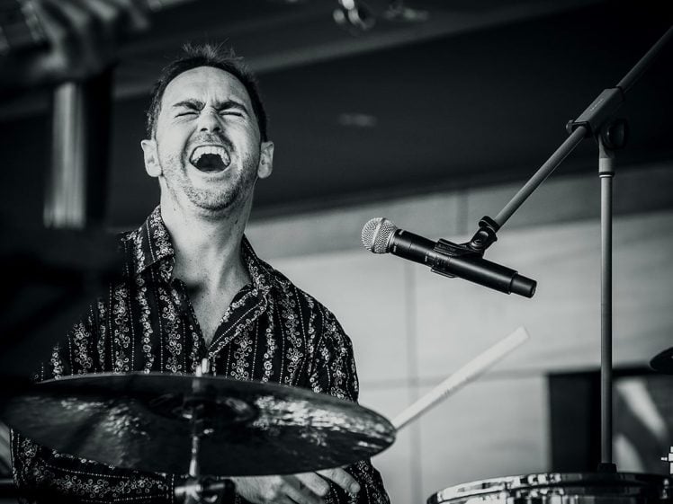 Image of Black and white photo of a man passionately playing drums, eyes closed and mouth wide open as if singing or shouting, with a microphone positioned near his face.