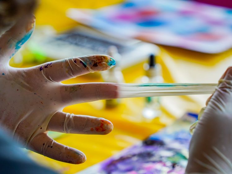 Image of A close-up of gloved hands stretching a piece of colourful slime, with splatters of paint on the gloves. Art supplies and abstract paintings are blurred in the background on a yellow surface.