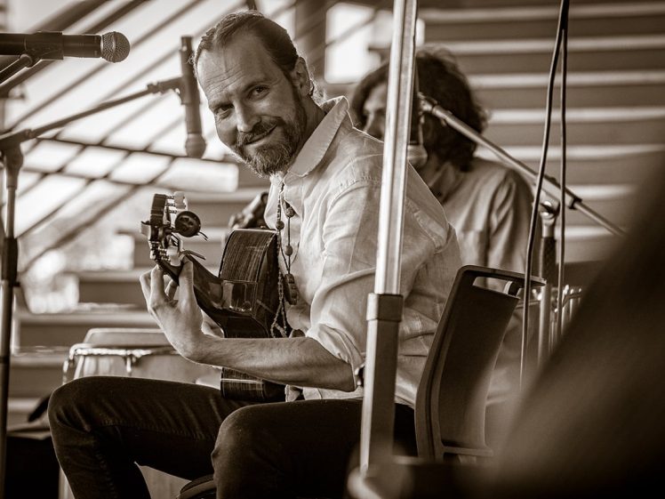 Image of A bearded man in a white shirt sits on a chair, playing an acoustic guitar and smiling at the camera. He is onstage with a microphone nearby, and another person is visible in the background. The image is in sepia tone.