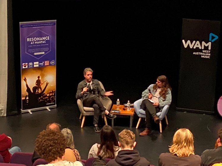 Image of Two people sit on stage having a discussion, with microphones, in front of an audience. Behind them are banners for Resonance at MANPAC and West Australian Music (WAM). A small table with water bottles sits between them.