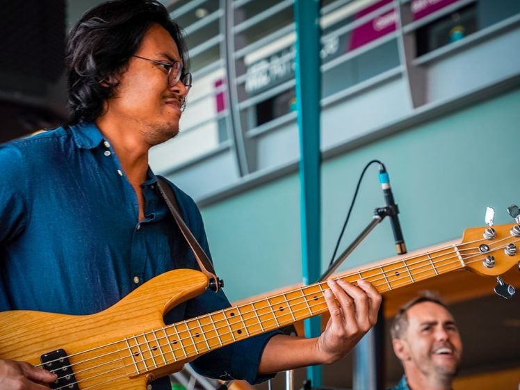 Image of A musician in a blue shirt plays a wooden electric bass guitar on stage while another person, slightly out of focus, smiles in the background. Microphones and a modern building are visible behind them.