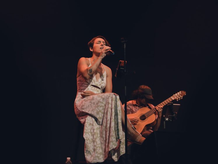 Image of A woman in a long, light-patterned dress sits on a stool singing into a microphone on stage; a man beside her plays an acoustic guitar. The background is dark, focussing attention on the performers.