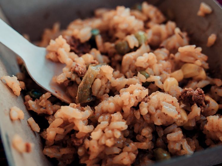 Image of Close-up of a cardboard carton filled with seasoned rice, minced meat, and chopped vegetables, with a plastic fork partially immersed in the food.