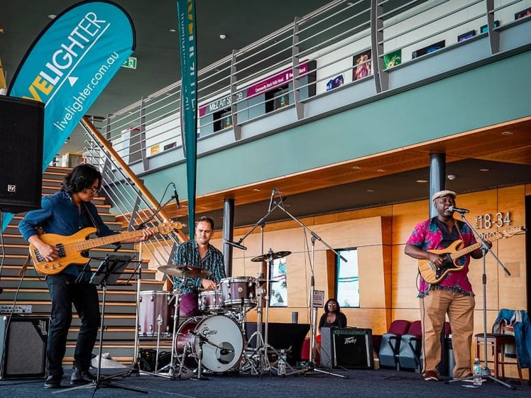 Image of A three-piece band performs indoors on a stage; the bassist, drummer, and guitarist/singer play under a “LiveLighter” banner. Stairs and spectators are visible in the modern venue.