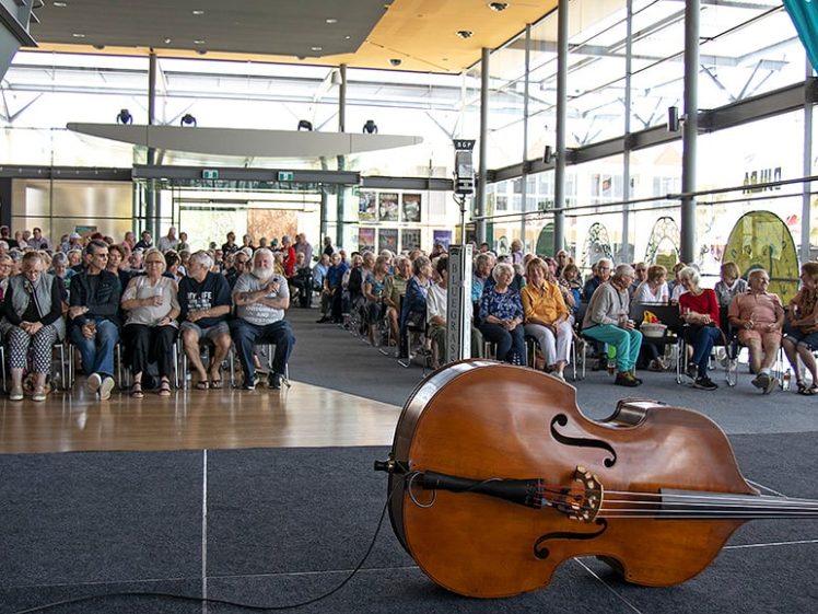 Image of A large audience sits in rows facing a stage inside a modern, glass-walled venue. A double bass rests on its side at the front of the empty stage, ready for a performance.