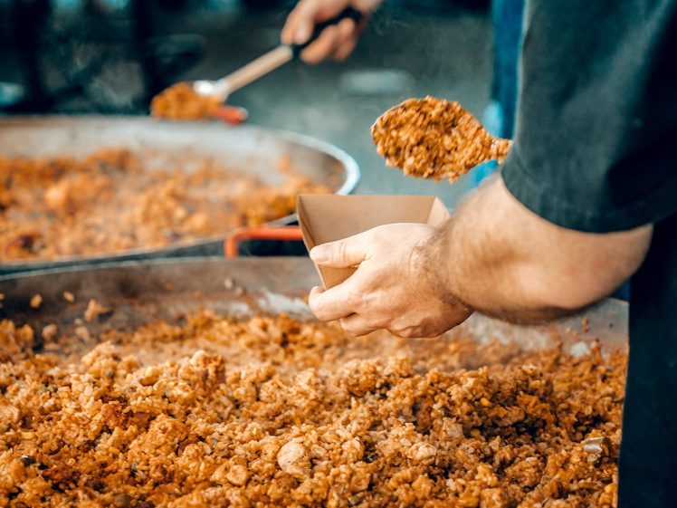 Image of A person spoons a serving of cooked rice from a large pan into a small brown paper container, with another person stirring food in the background.