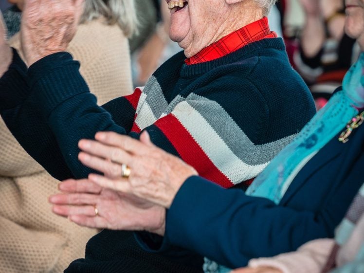 Image of An elderly man with white hair, wearing a striped jumper, smiles and claps whilst sitting among a group of older adults at an indoor event.