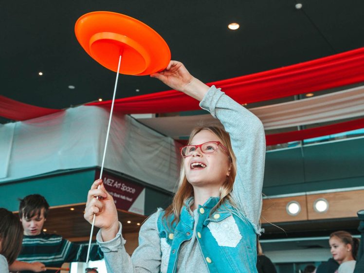 Image of A smiling girl with glasses spins an orange plate on a stick indoors, surrounded by other children. Red and white fabric drapes hang from the ceiling in the background.
